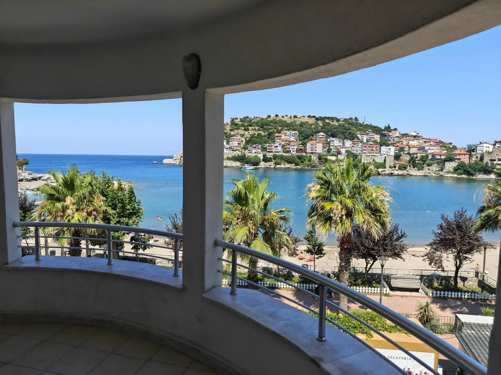 Aile odasının kavisli balkonundan Amasra koyu: palmiyeler, sahil promenadı ve karşı yamaçtaki evler