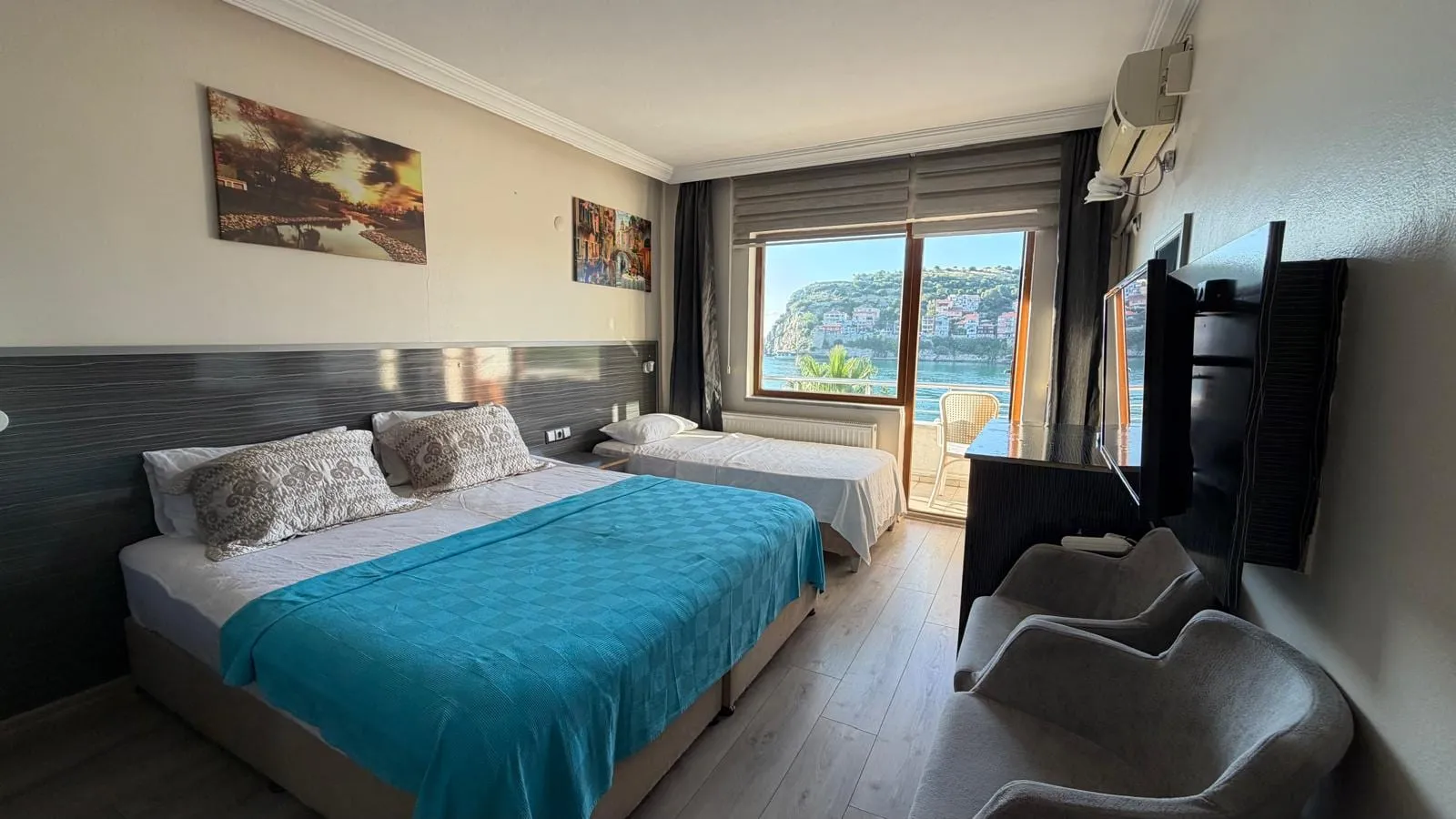 Aile odası: mavi örtülü çift yatak, ilave yatak, oturma koltukları ve denize bakan balkon kapısı