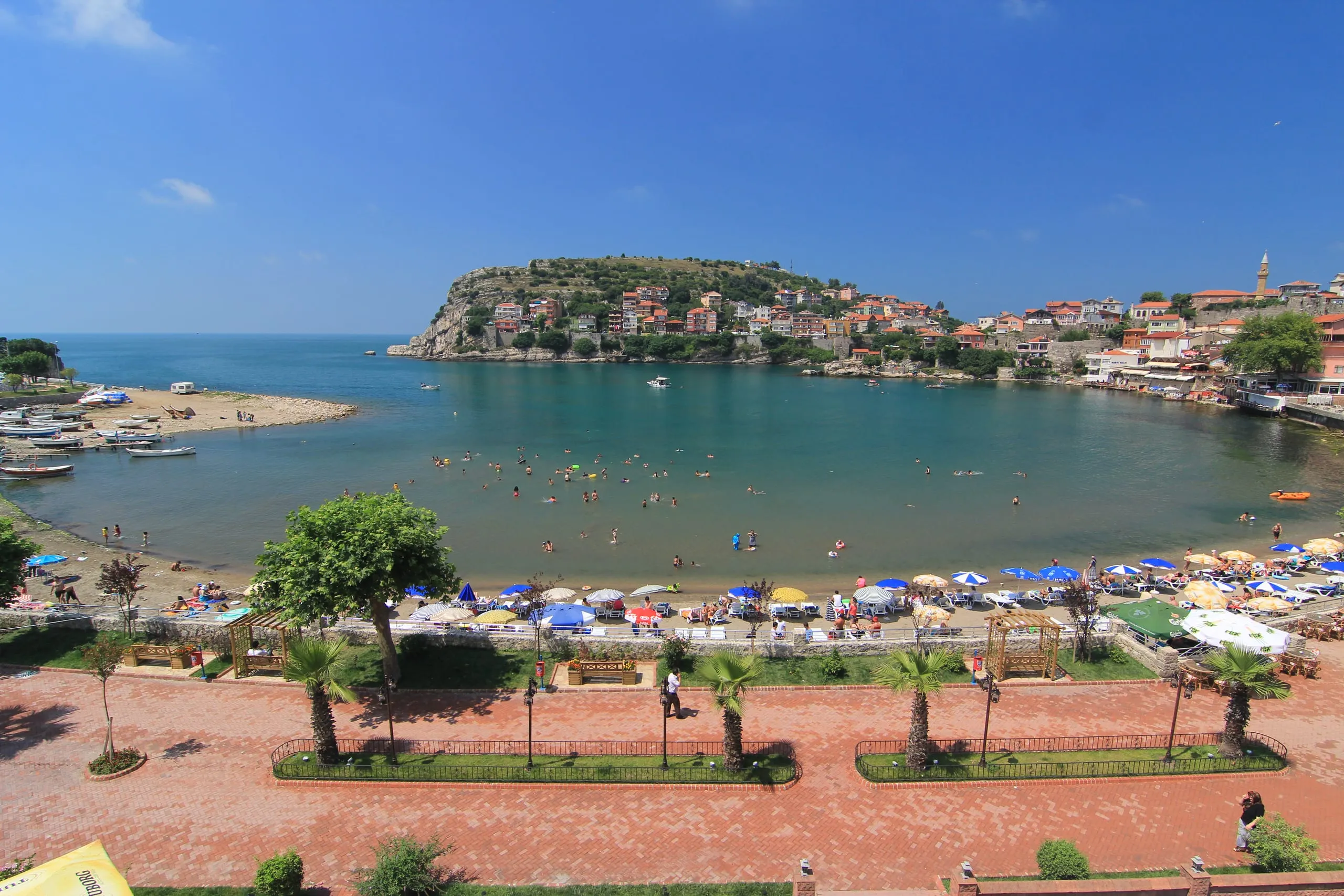 Amasra Büyük Liman koyu: kumsal, şemsiyeli sahil promenadı ve karşıda yarımadadaki tarihi Amasra evleri