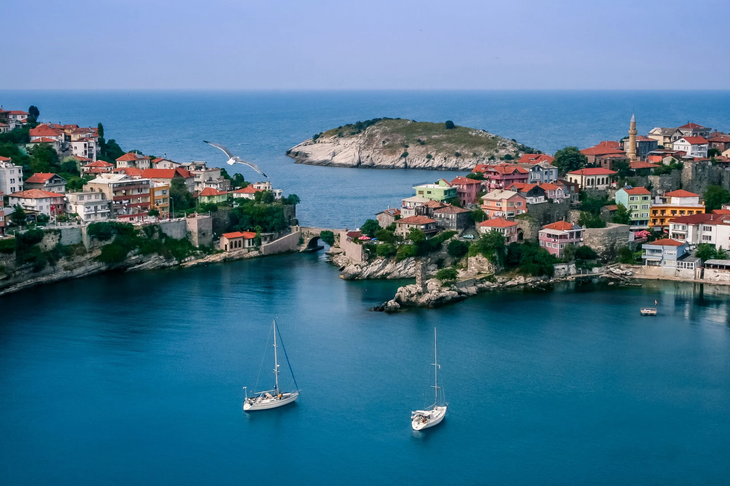 Amasra'da taş kemer köprü, kale surları ve karşıdaki ada; koyda demirlemiş iki yelkenli ve uçan bir martı