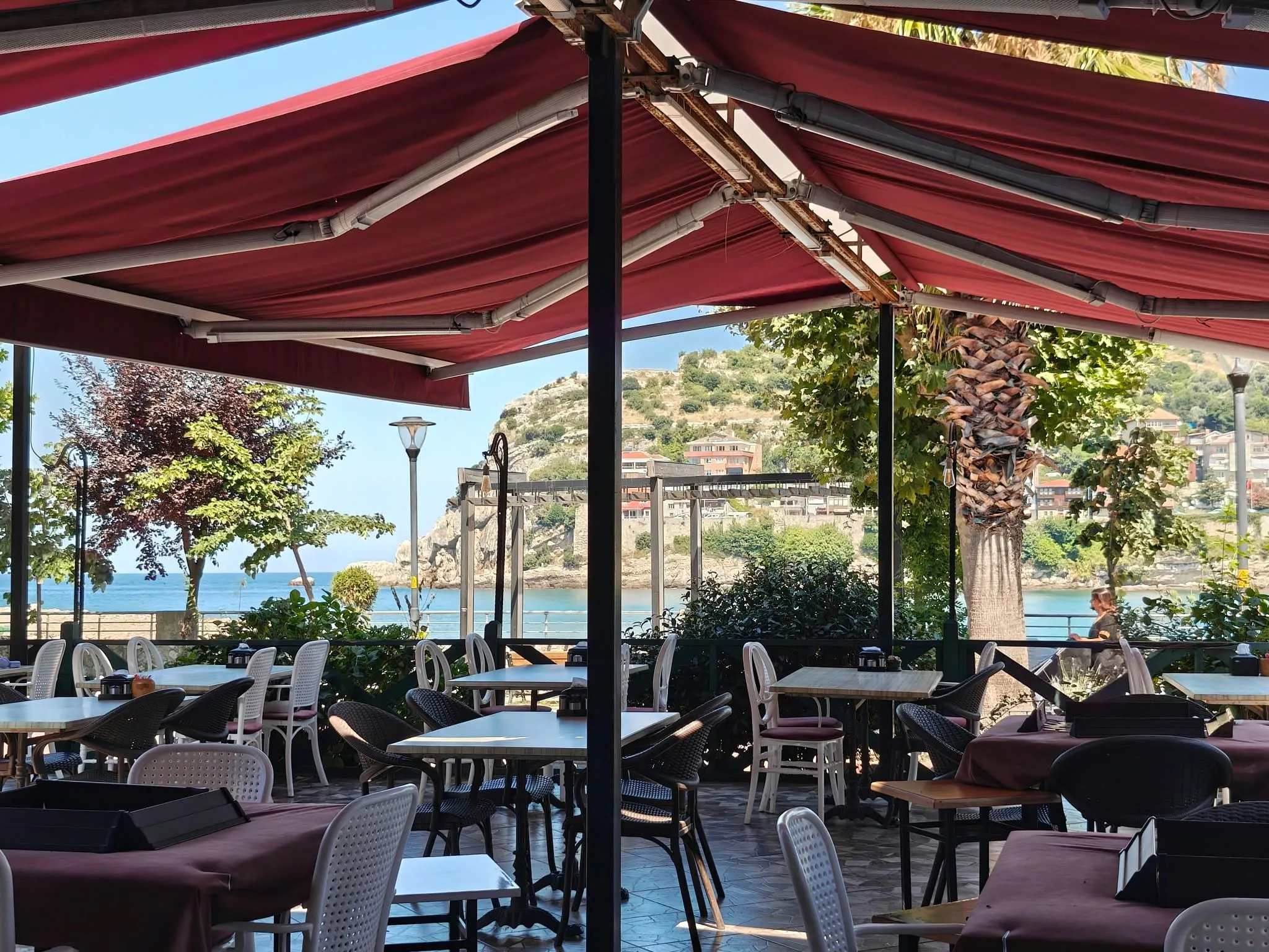 Cafe terası: bordo tenteler altında masalar ve arkada Amasra yarımadası ile deniz