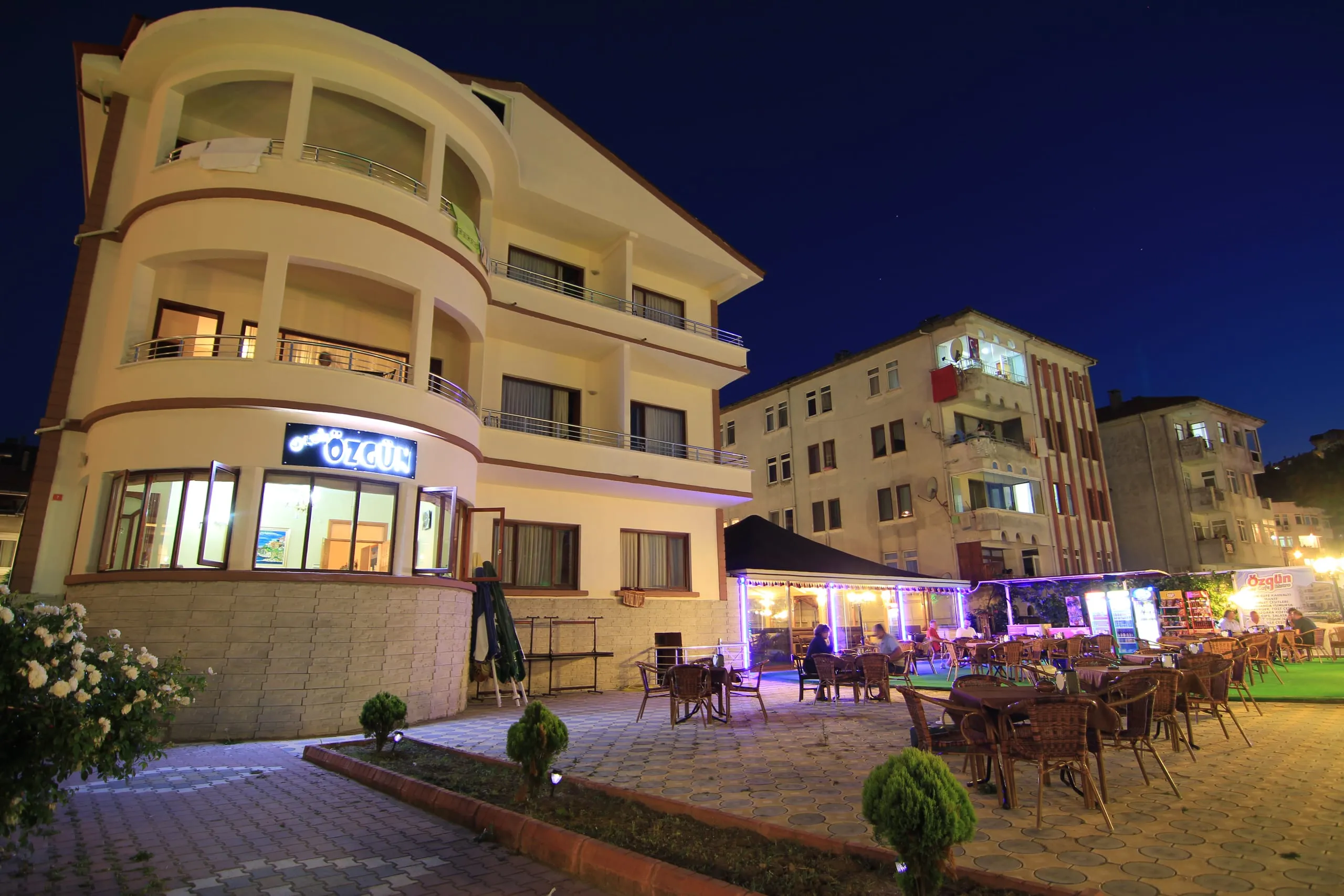 Amasra Özgün Otel'in akşam saatlerinde aydınlatılmış cephesi; balkonlu üç katlı bina ve girişindeki Özgün Cafe terası